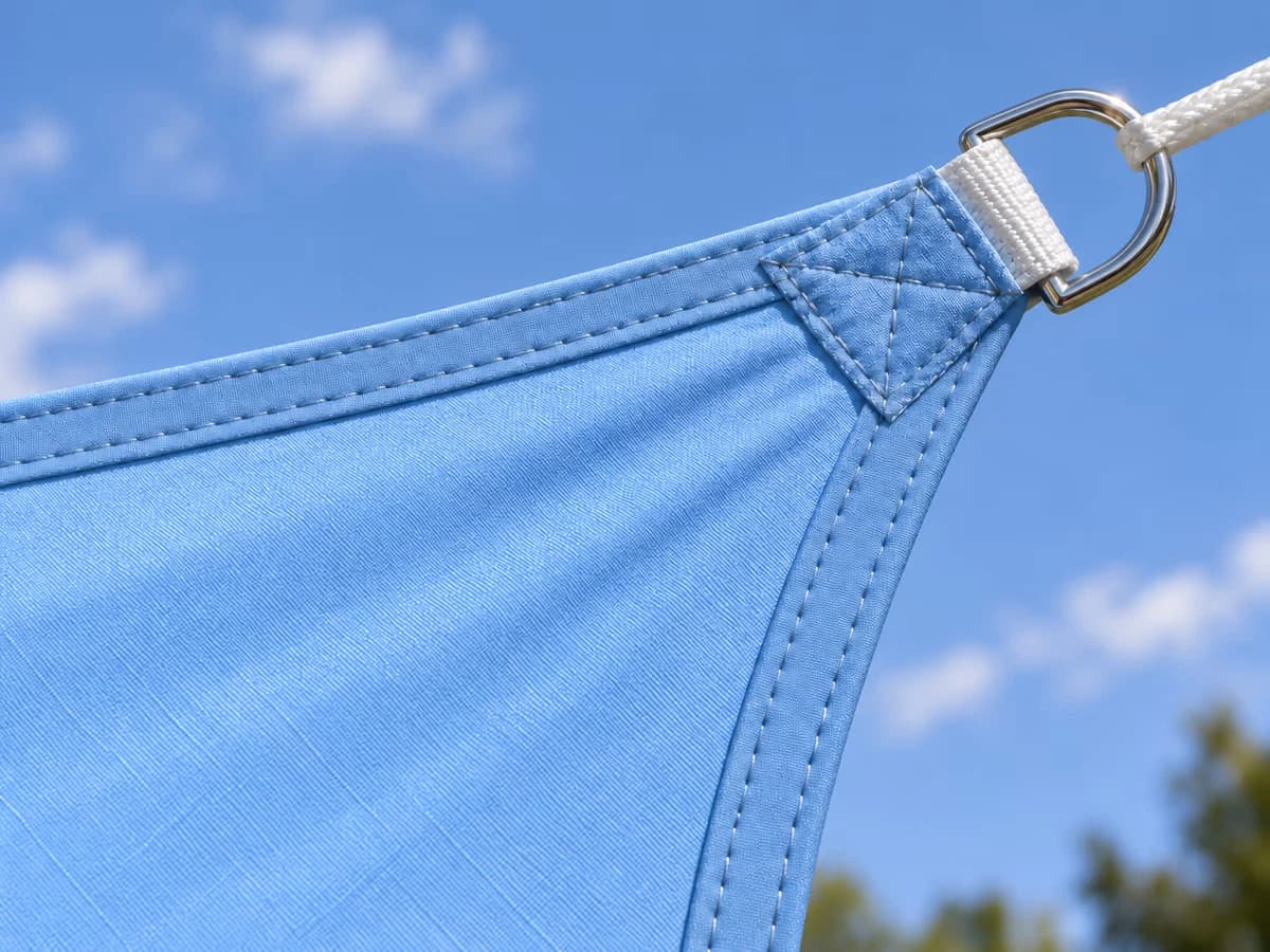 Gros plan sur le bord arqué d'une voile d'ombrage bleu ciel et sur l'anneau en D cousu dans la sangle d'une pointe, toile tendue