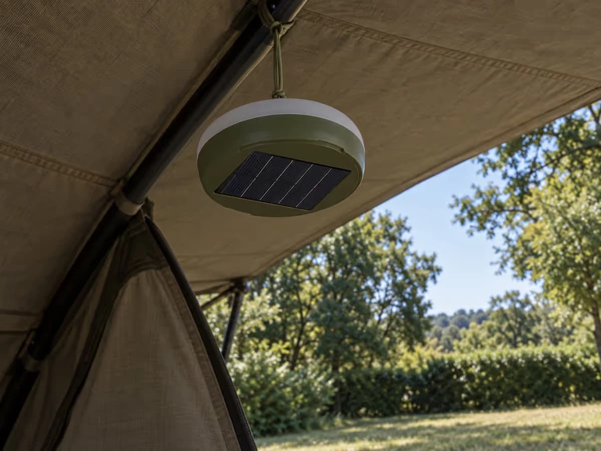 Boîtier solaire suspendu à l'ombre sous une toile de tente, panneau bleu visible au dos, arbres en arrière-plan