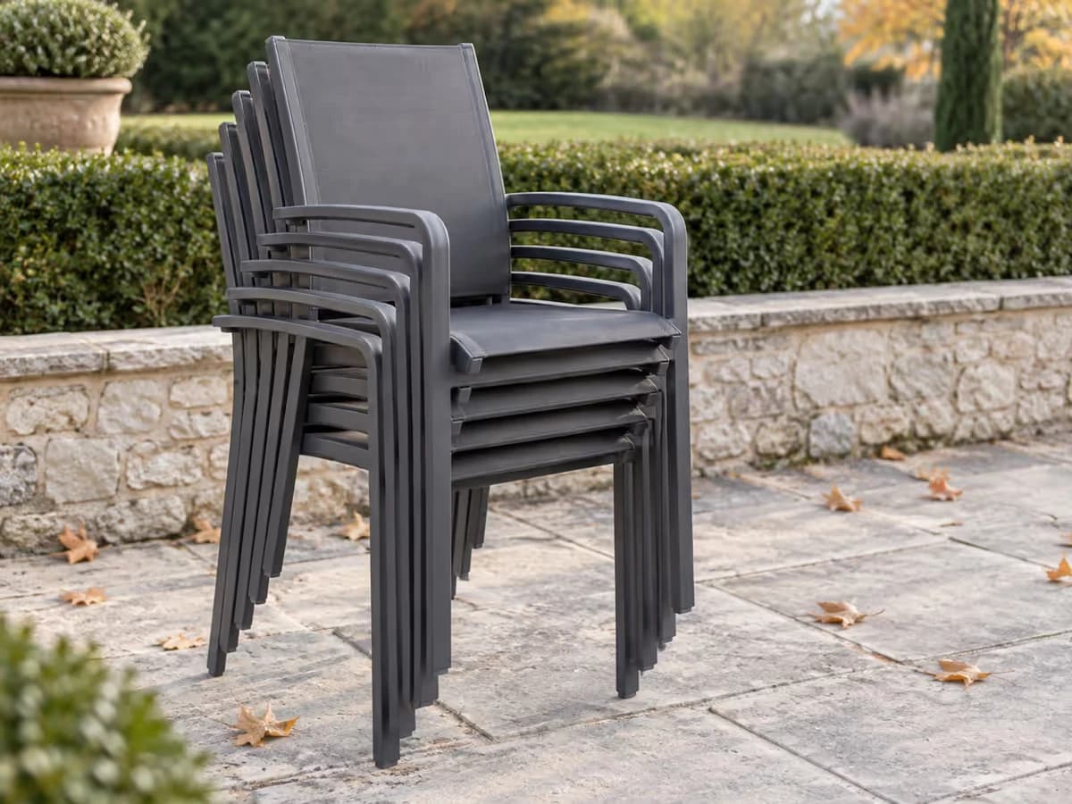 Pile de chaises de jardin empilées contre un mur de terrasse, dossiers alignés et assises emboîtées, sur dalles claires un matin sec d'automne