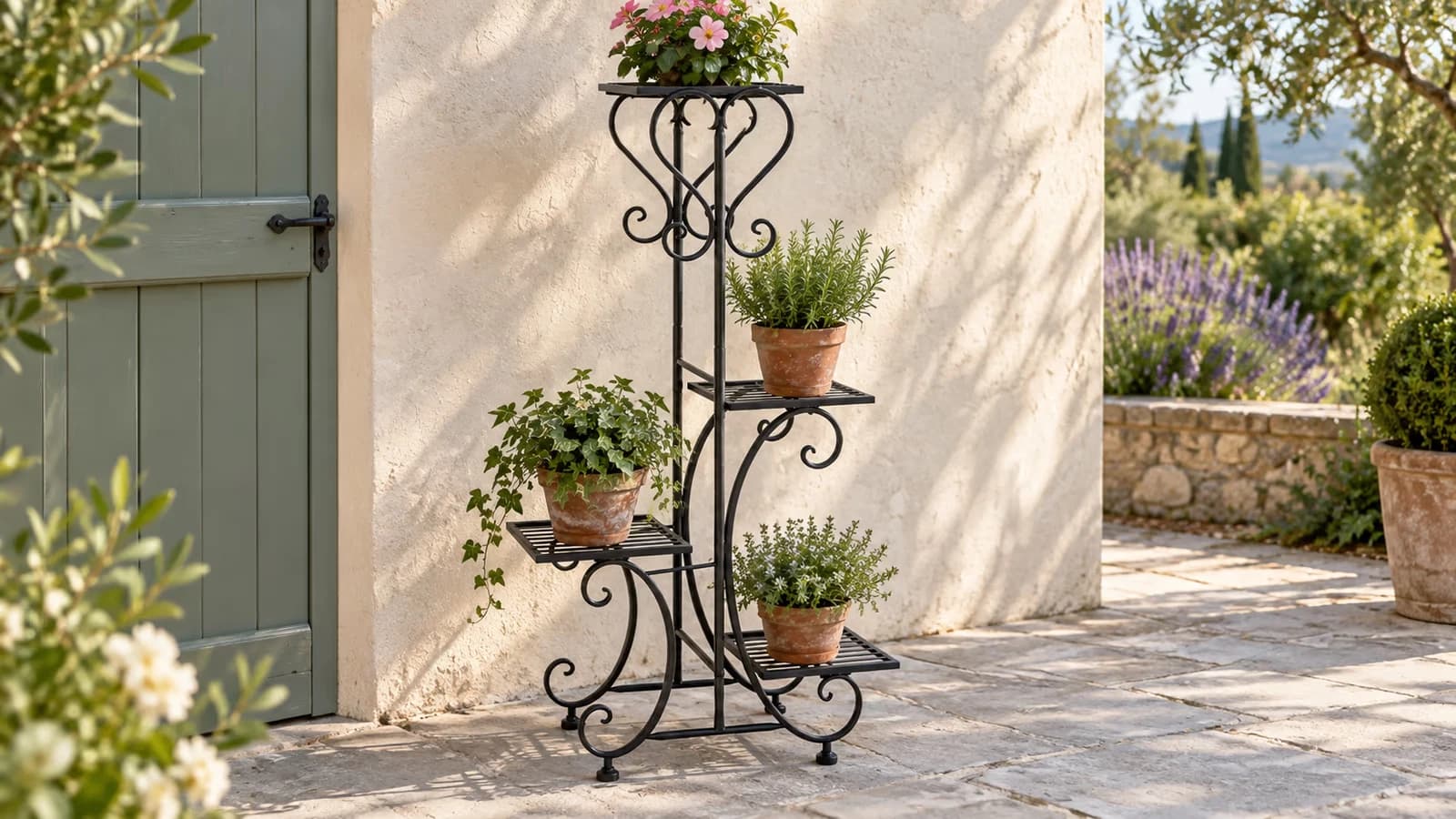 Porte plantes en fer forgé à quatre plateaux posé contre un mur de terrasse, quatre petits pots de fleurs en place