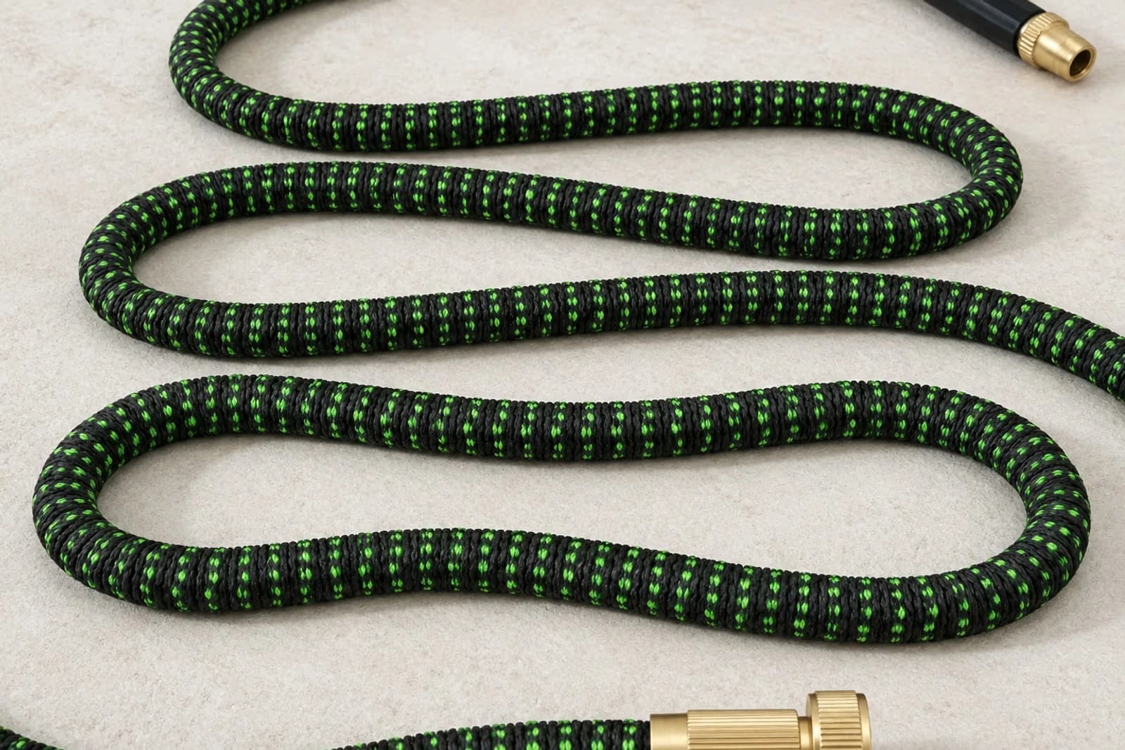 Tuyau extensible déployé à plat sur fond studio off-white, gaine polyester verte tissée visible texture, raccords laiton brillants aux deux extrémités