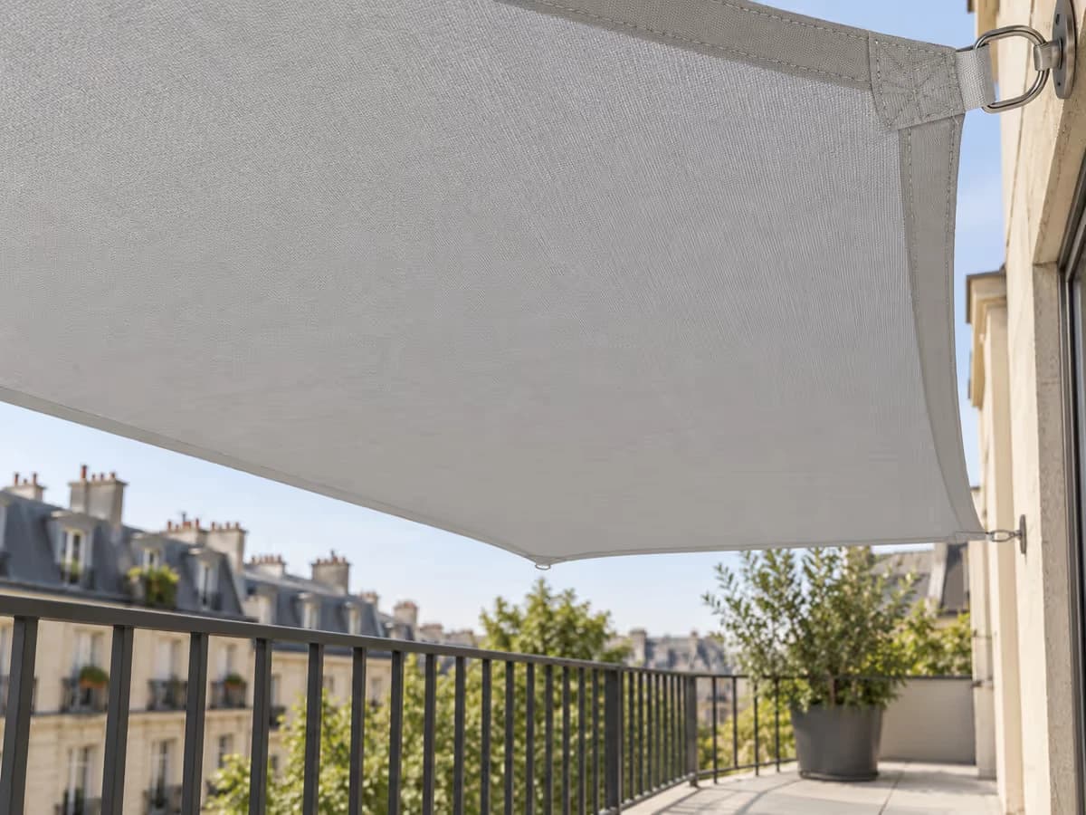 Voile d'ombrage grise tendue sans pli au-dessus d'un balcon, vue de dessous, anneaux et sangle visibles sur le bord