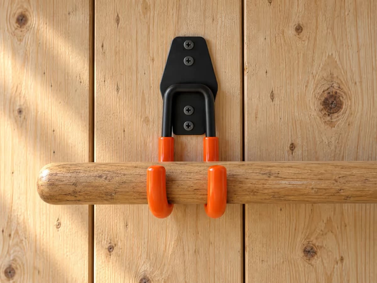 Manche de bêche en bois posé au tiers bas entre les deux bras orange d'un crochet mural, sur un mur d'abri de jardin