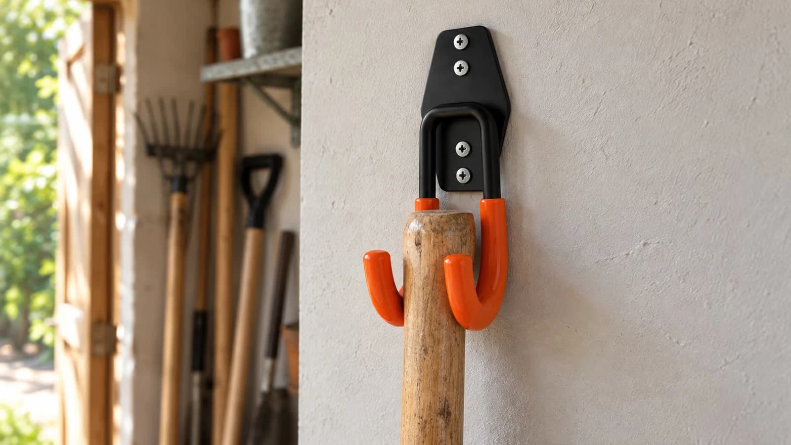 Crochet mural en U en fer noir fixé sur un mur de garage, un manche de bêche suspendu entre les deux bras gainés d'orange