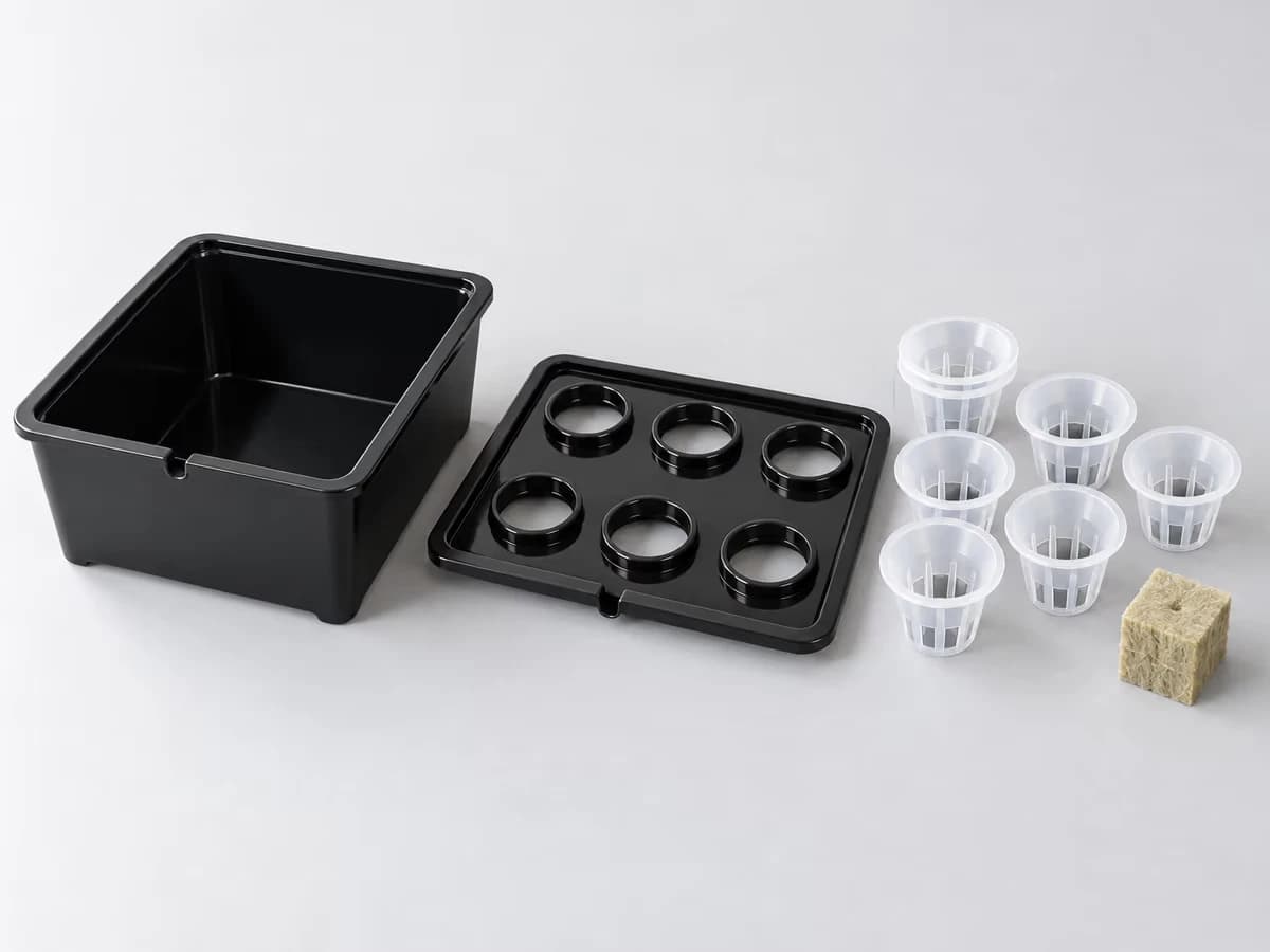 Trois pièces d'un kit de germination alignées sur un fond clair : bac noir, couvercle percé de six ouvertures, six paniers en maille translucide, un cube de laine de roche posé à côté d'eux