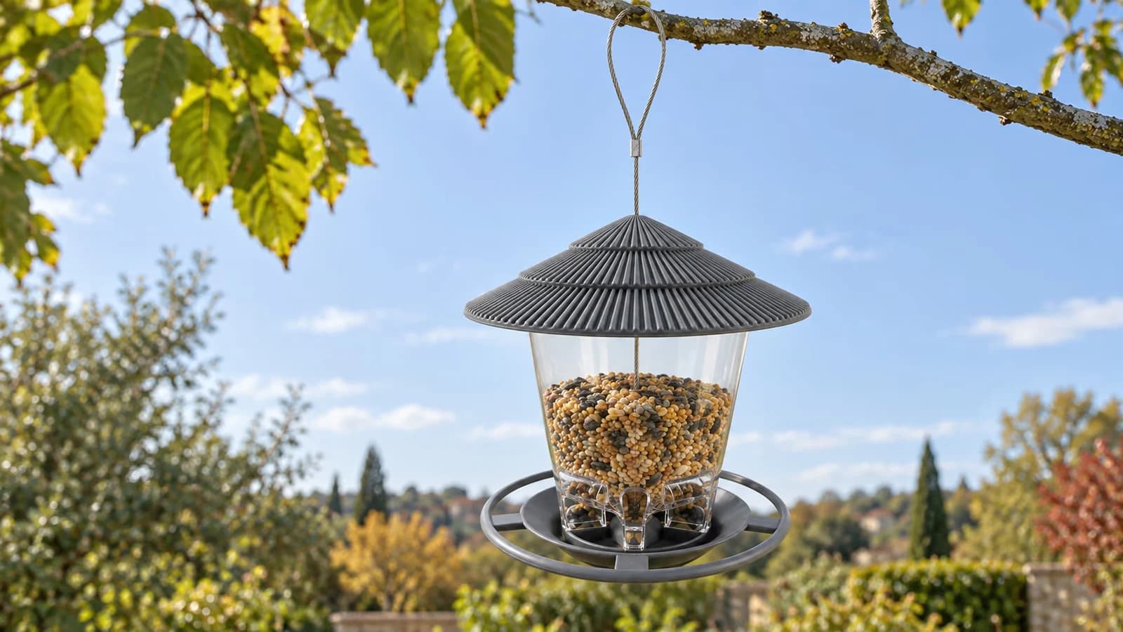 Mangeoire à suspendre gris anthracite au toit conique à deux niveaux, suspendue par une boucle de câble à une branche nue, ciel d'automne dégagé