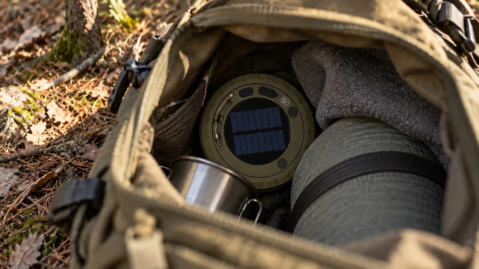 Lampe ronde de camping posée face contre terre au fond d'un sac de randonnée ouvert, en fin d'après-midi sous des arbres