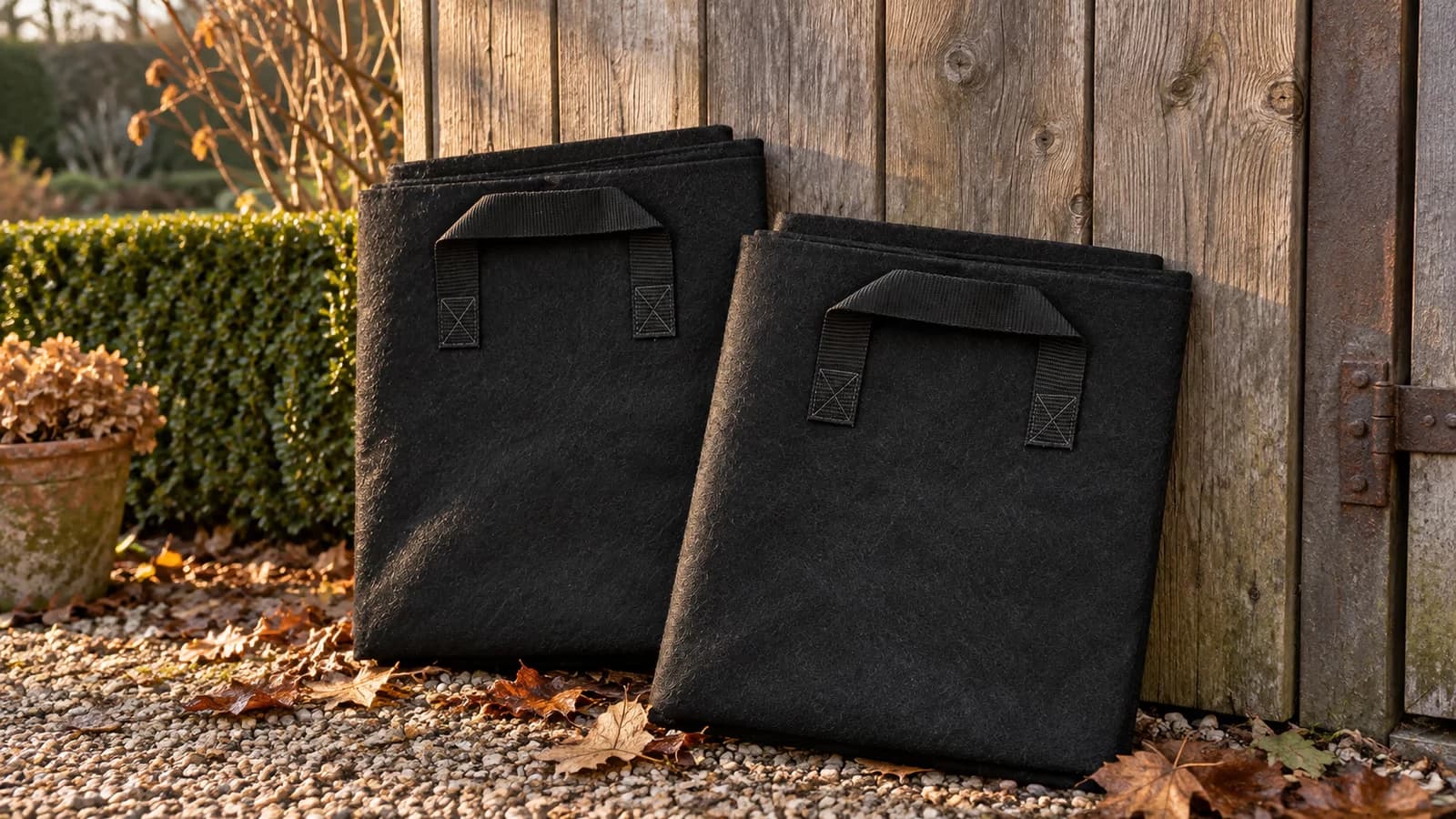 Deux sacs de culture en textile noir, vides et pliés à plat contre un mur de cabanon, lumière rasante de fin d'automne