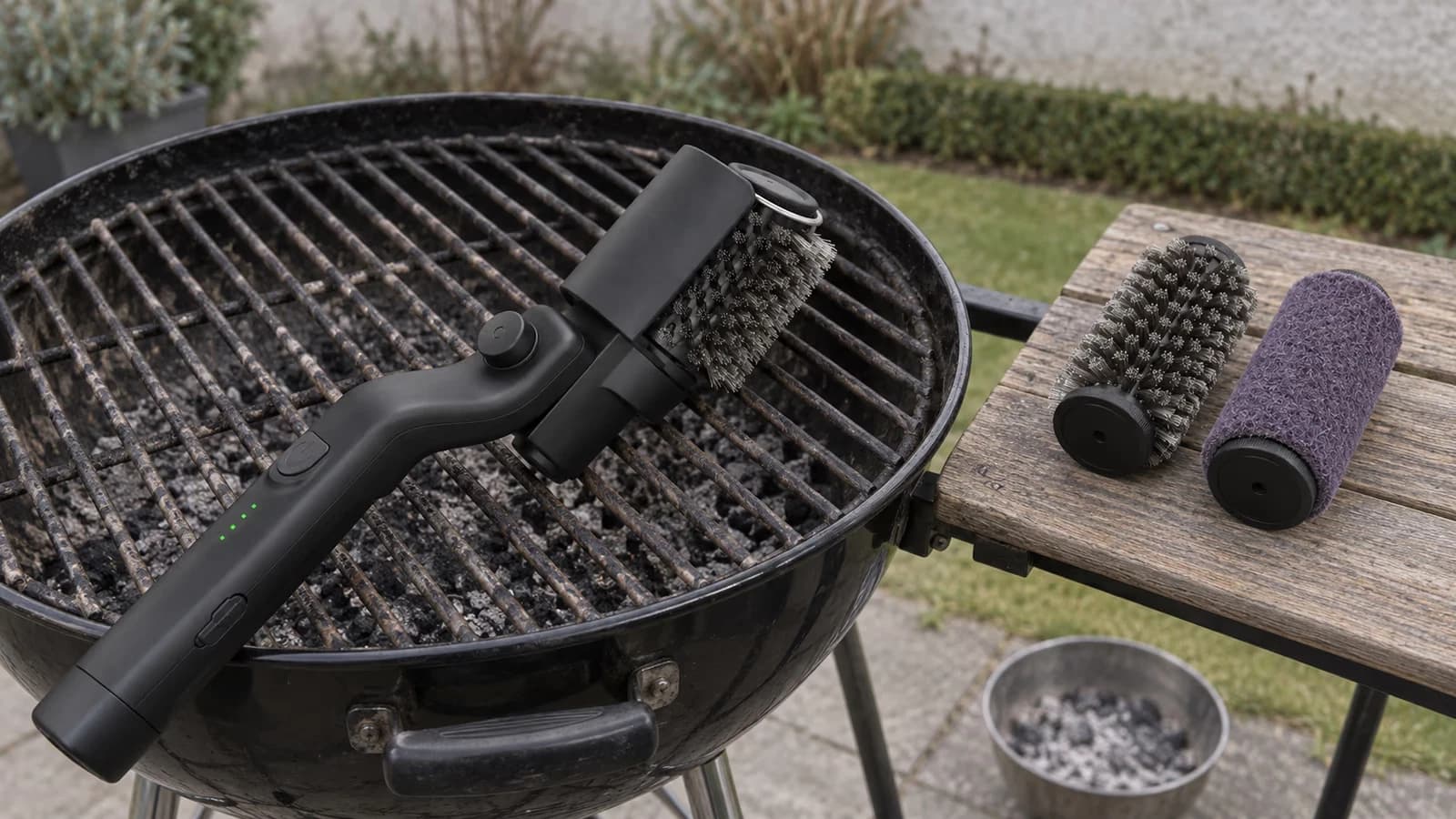 Brosse électrique sans fil posée sur la grille d'un barbecue, deux têtes détachables alignées à côté sur une planche