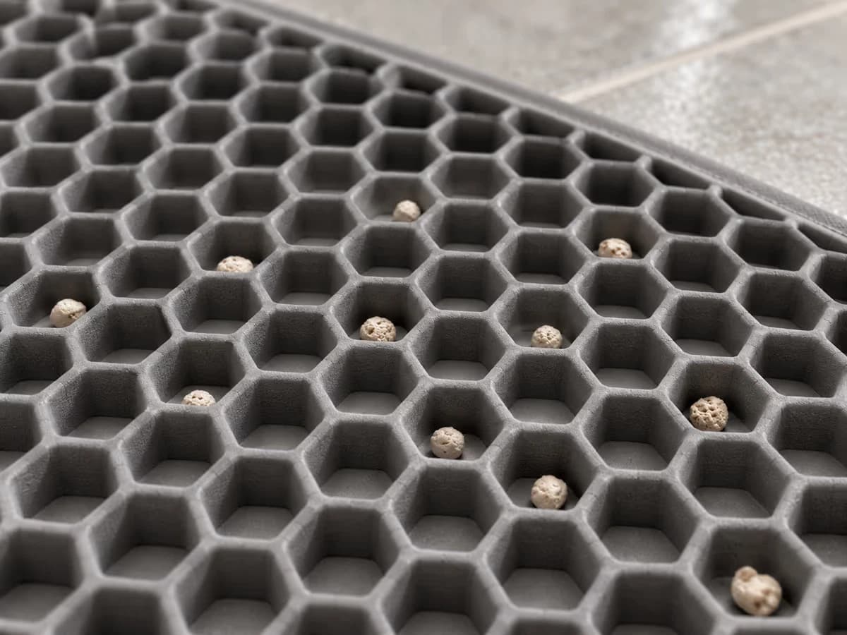 Gros plan sur les alvéoles hexagonales d'un tapis de litière gris, granulés d'argile retenus entre les reliefs, carrelage clair en arrière-plan