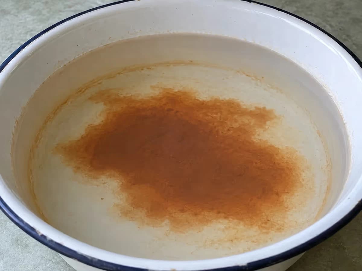 Tache brun-rouge de sédiment fin déposée au fond d'une bassine émaillée blanche, avec une auréole ocre pâle sur l'émail