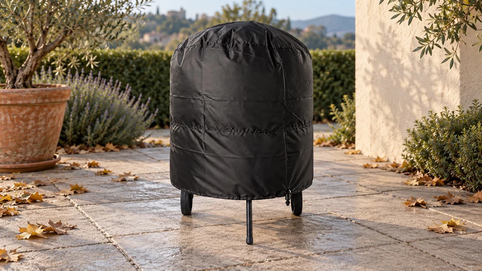 Gril à charbon rond recouvert de sa couverture noire, cordon serré à la base de la cuve, sur une terrasse en dalles un matin d'automne