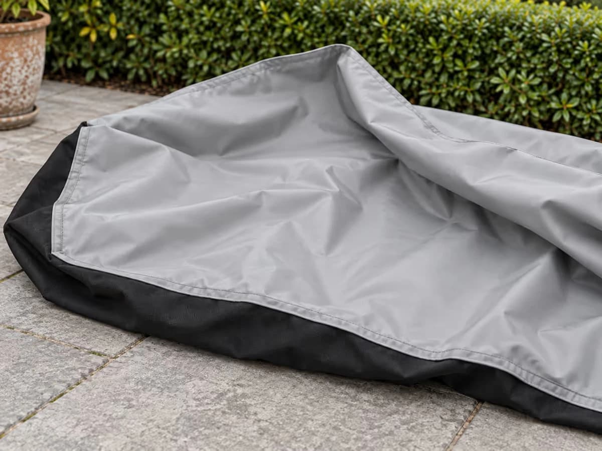 Doublure PVC gris clair d'une bâche de table de jardin retournée sur des dalles sèches, couture et ourlet visibles, face extérieure noire au sol