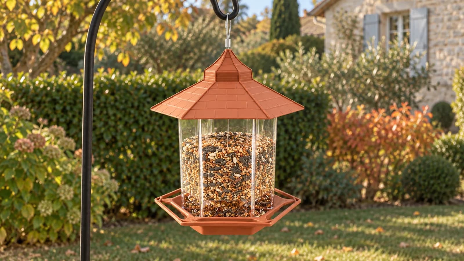 Mangeoire à silo hexagonale au toit de tuiles terracotta suspendue à une potence métallique, jardin d'automne au fond