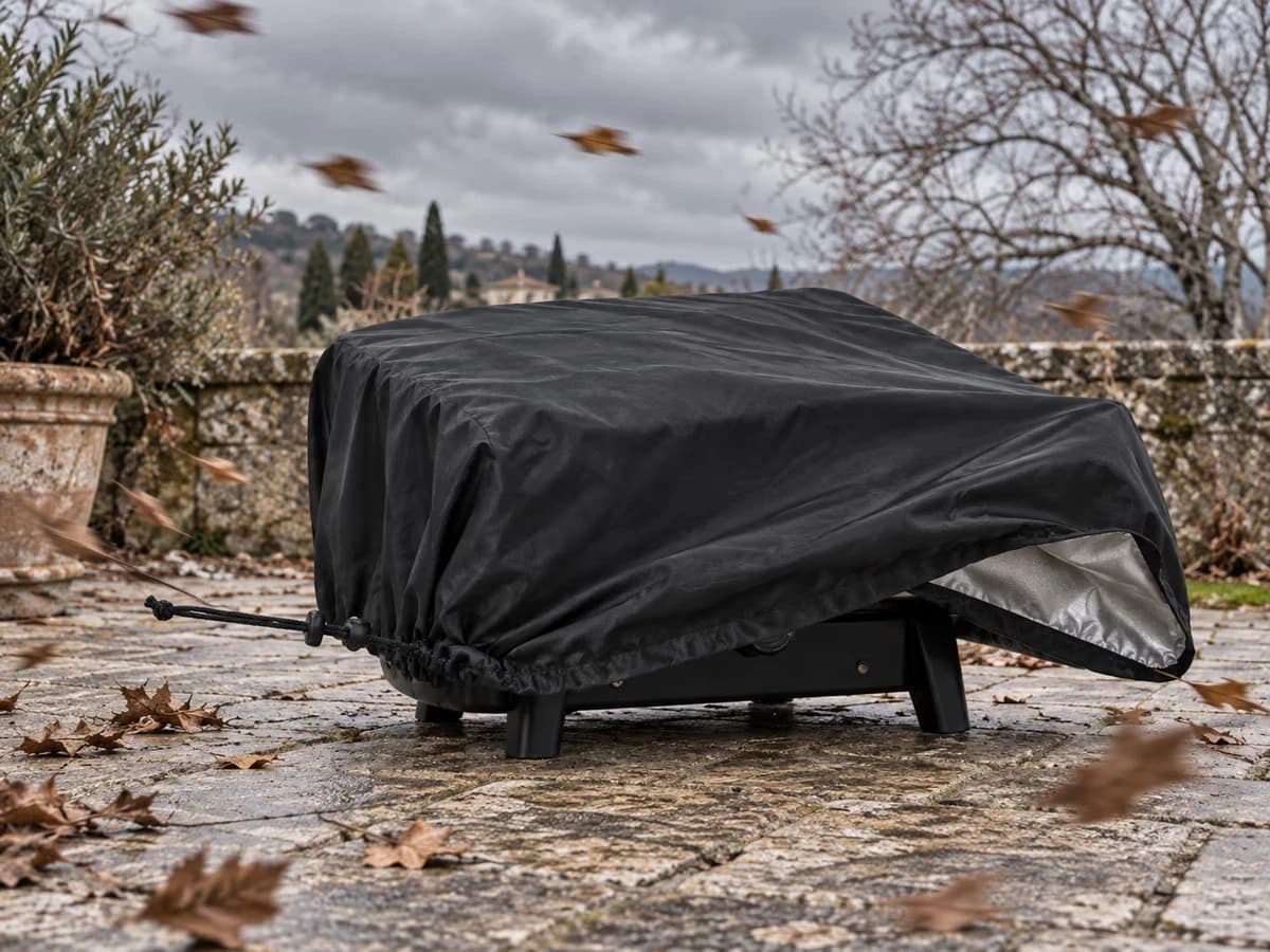 Housse noire soulevée par une rafale sur un appareil de cuisson de table, ourlet décollé du sol, terrasse balayée par le vent