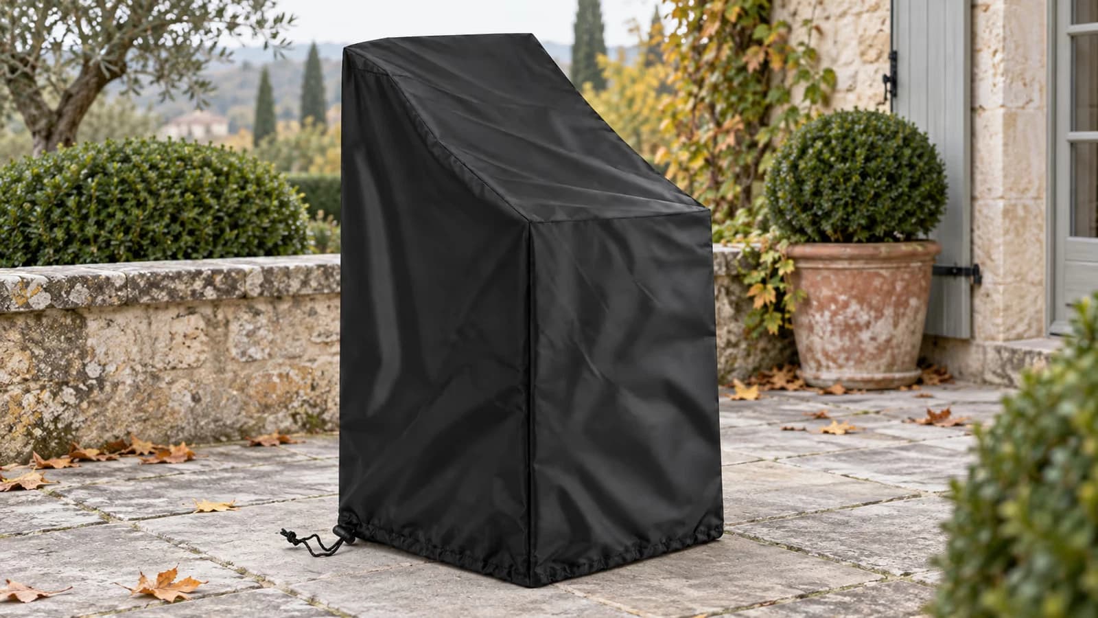 Housse de protection noire posée sur une pile de chaises de jardin empilables, cordon serré au pied, sur une terrasse en dalles en fin de saison