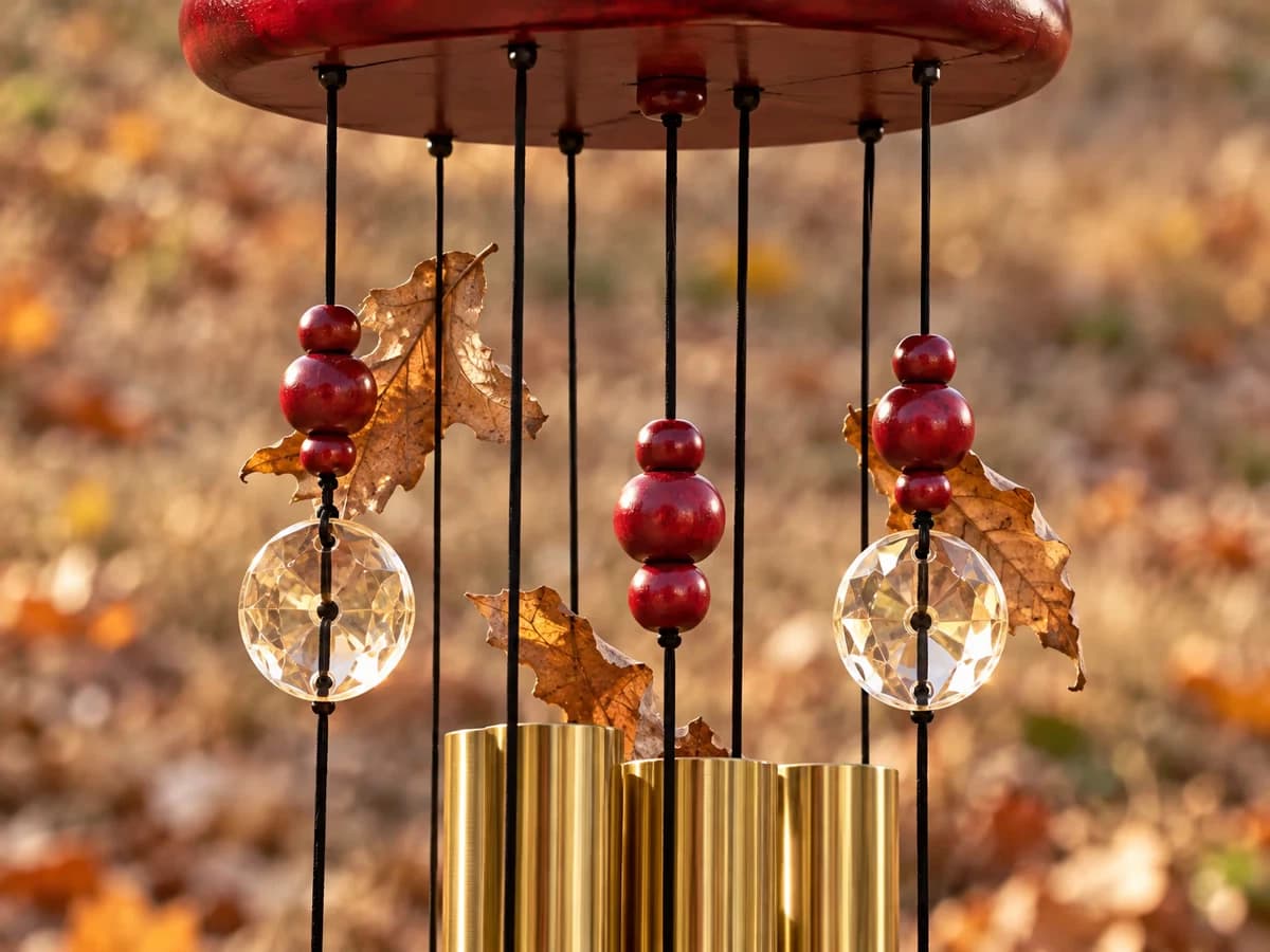 Gros plan sur les suspentes d'un carillon doré, perles rouges et disques de cristal soulevés une à une, feuilles mortes au sol