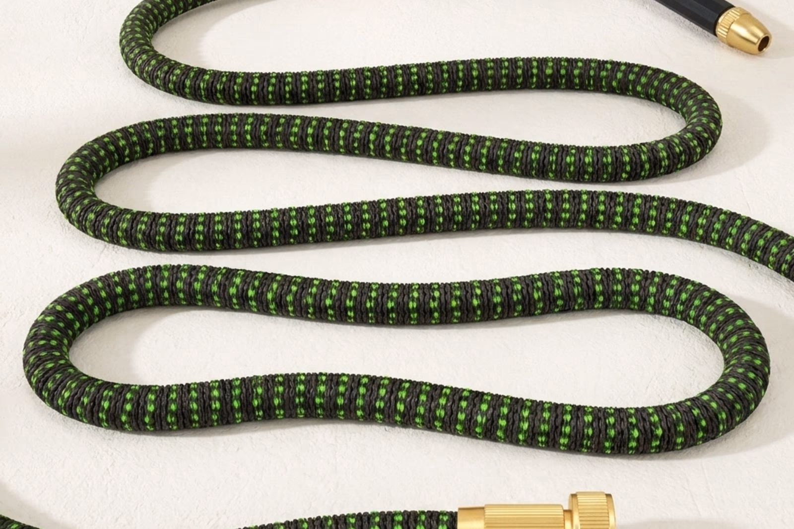 Tuyau extensible deploye a plat sur fond studio off-white, gaine polyester verte tissee visible texture, raccords laiton brillants aux deux extremites