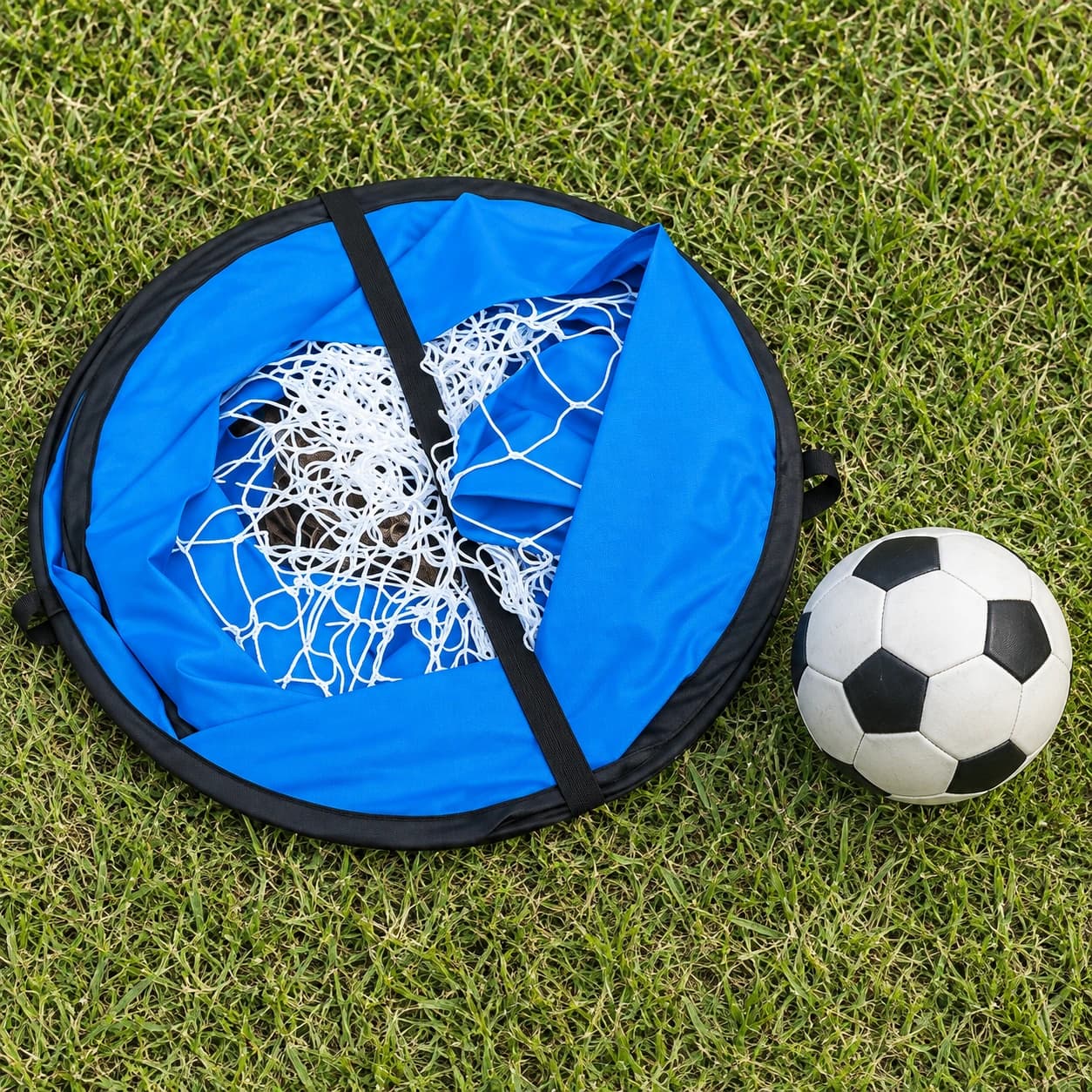 But de football pop-up enfant replié en disque bleu sur l'herbe, filet rangé et sangles croisées