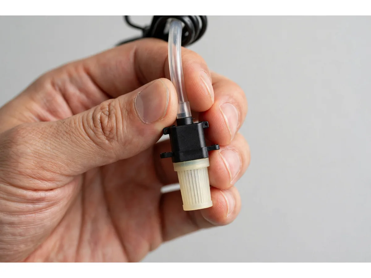 Une main adulte tient la pompe entre le pouce et l'index sur fond blanc, crépine crème et court tuyau transparent, taille d'une phalange de pouce