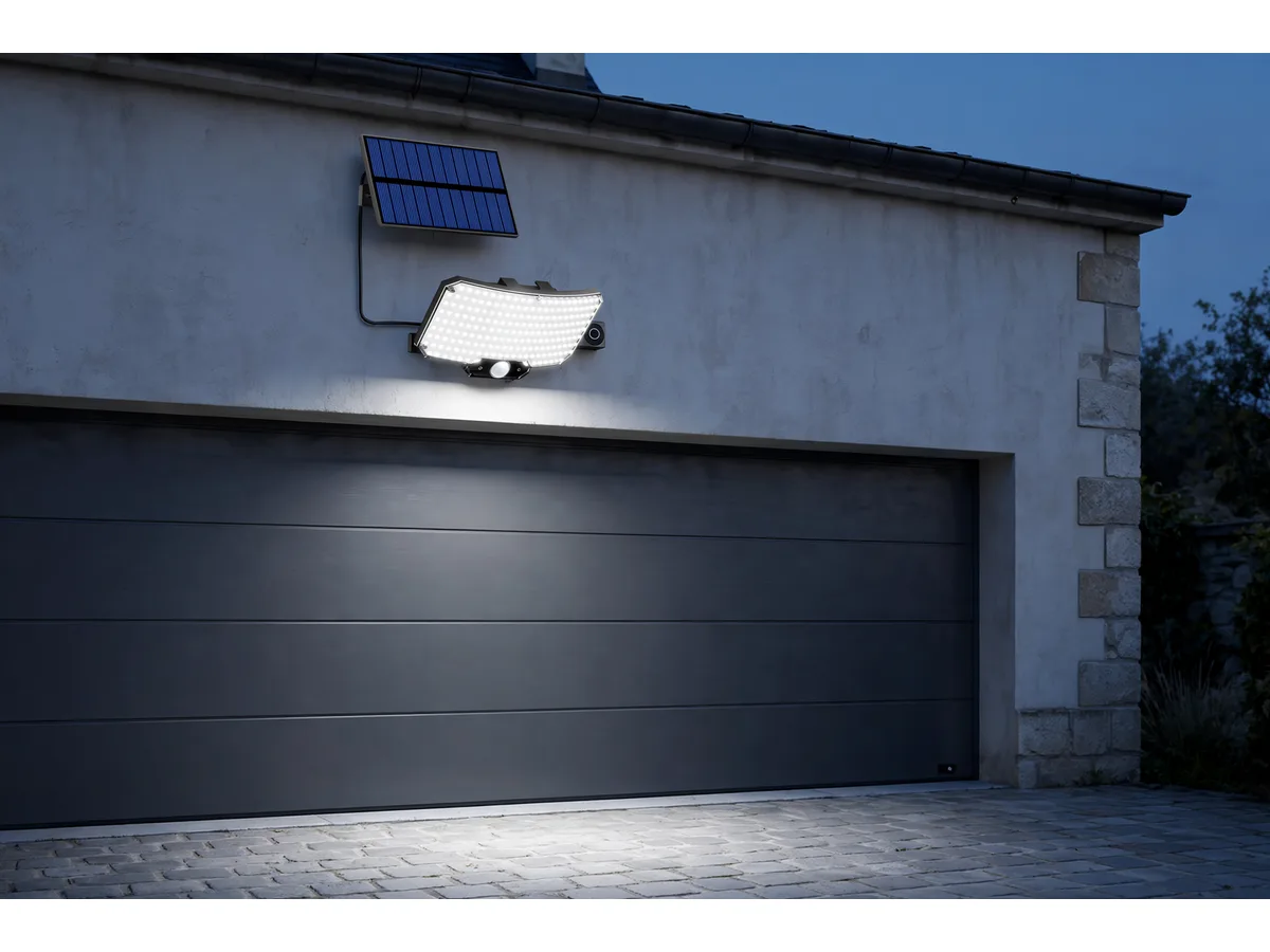De nuit, le bloc fixé en haut d'un mur de garage, panneau déporté plus haut sur le même mur, large nappe de lumière blanche sur la cour pavée et sur le portail
