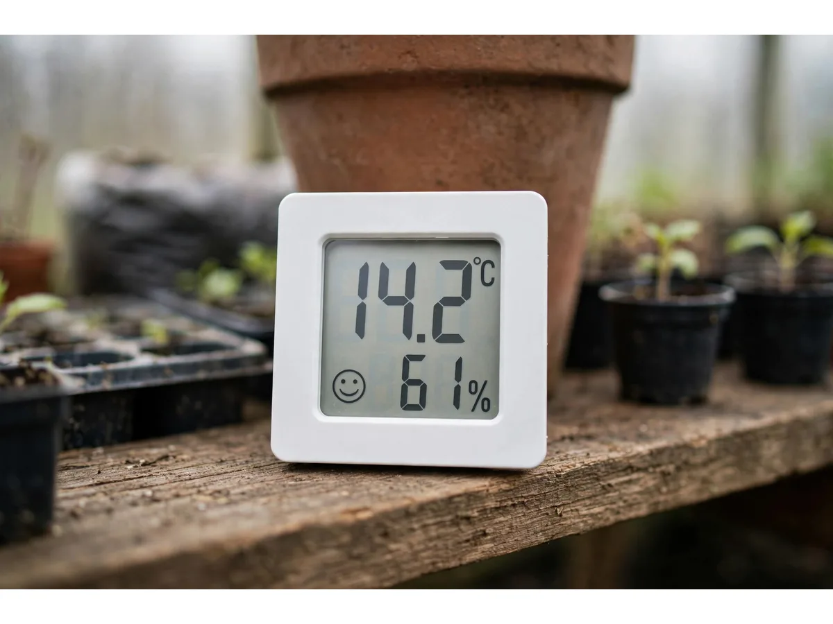 Le boîtier posé sur une étagère en bois d'une petite serre, contre un pot en terre cuite, godets de semis et jeunes pousses flous autour