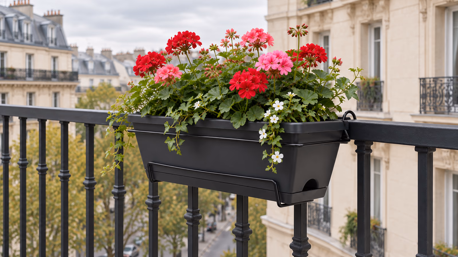 Jardinière à crochets garnie posée sur un garde-corps de balcon, plateau récupérateur en place sous le bac, immeuble en arrière-plan
