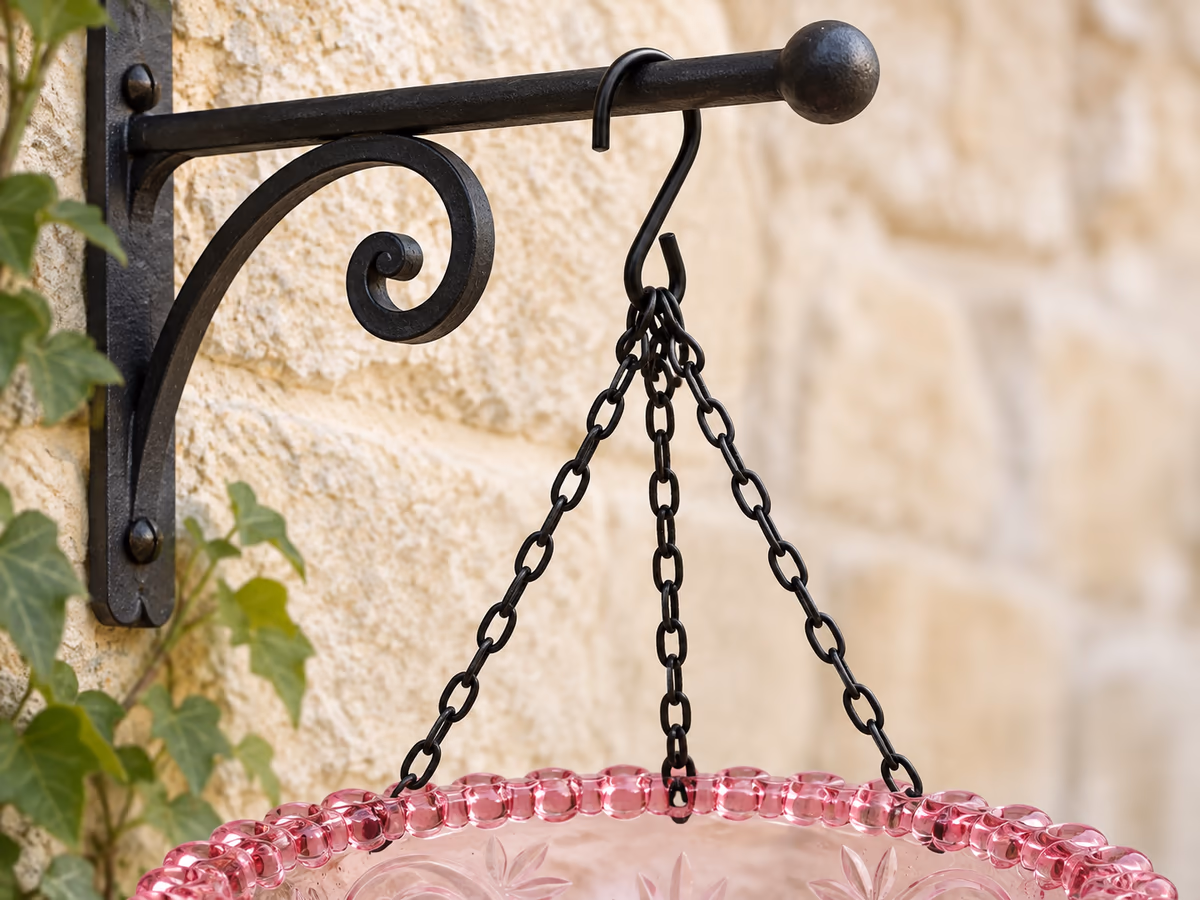 Détail d'un crochet ouvert passé sur le bras d'une potence de jardinière en fer forgé, avec la suspension raccourcie au plus près du support
