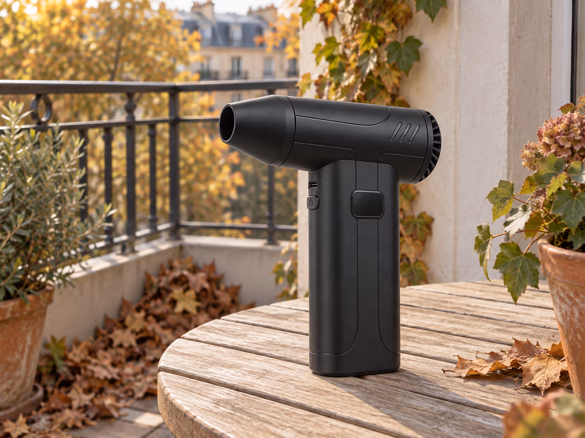 Mini souffleur rechargeable noir posé sur une table de balcon, feuilles mortes rassemblées dans l'angle du garde-corps en automne