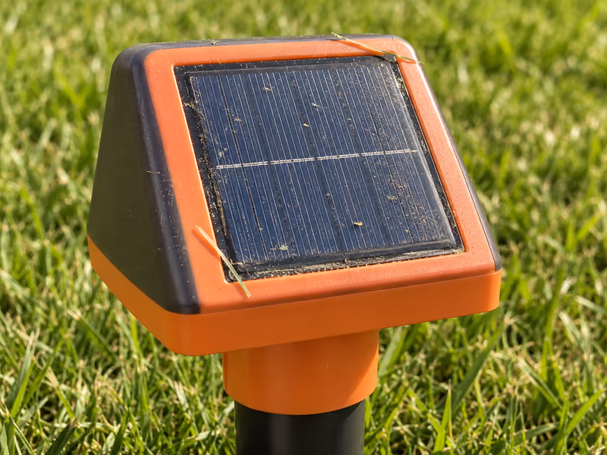 Face solaire inclinée d'un boîtier orange dans une pelouse, brins d'herbe tondue et poussière déposés sur le verre
