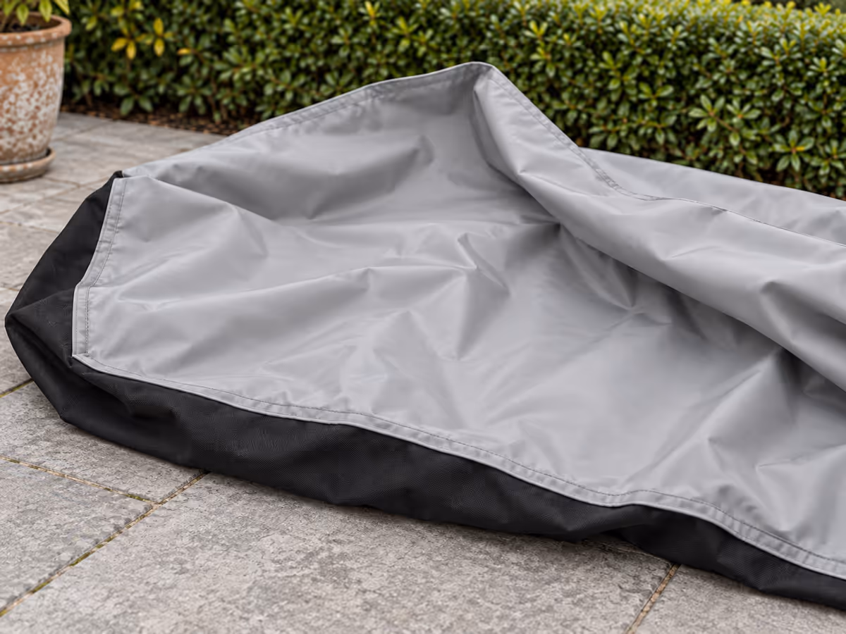 Doublure PVC gris clair d'une bâche de table de jardin retournée sur des dalles sèches, couture et ourlet visibles, face extérieure noire au sol