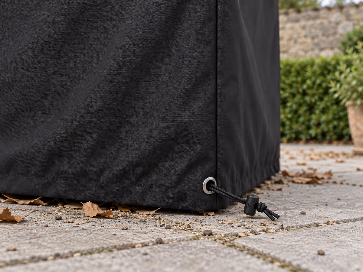Bas d'une bâche noire de table de jardin au niveau des dalles, cordon sorti par un œillet métallique et bloqueur serré contre la toile, feuilles mortes le long de la retombée