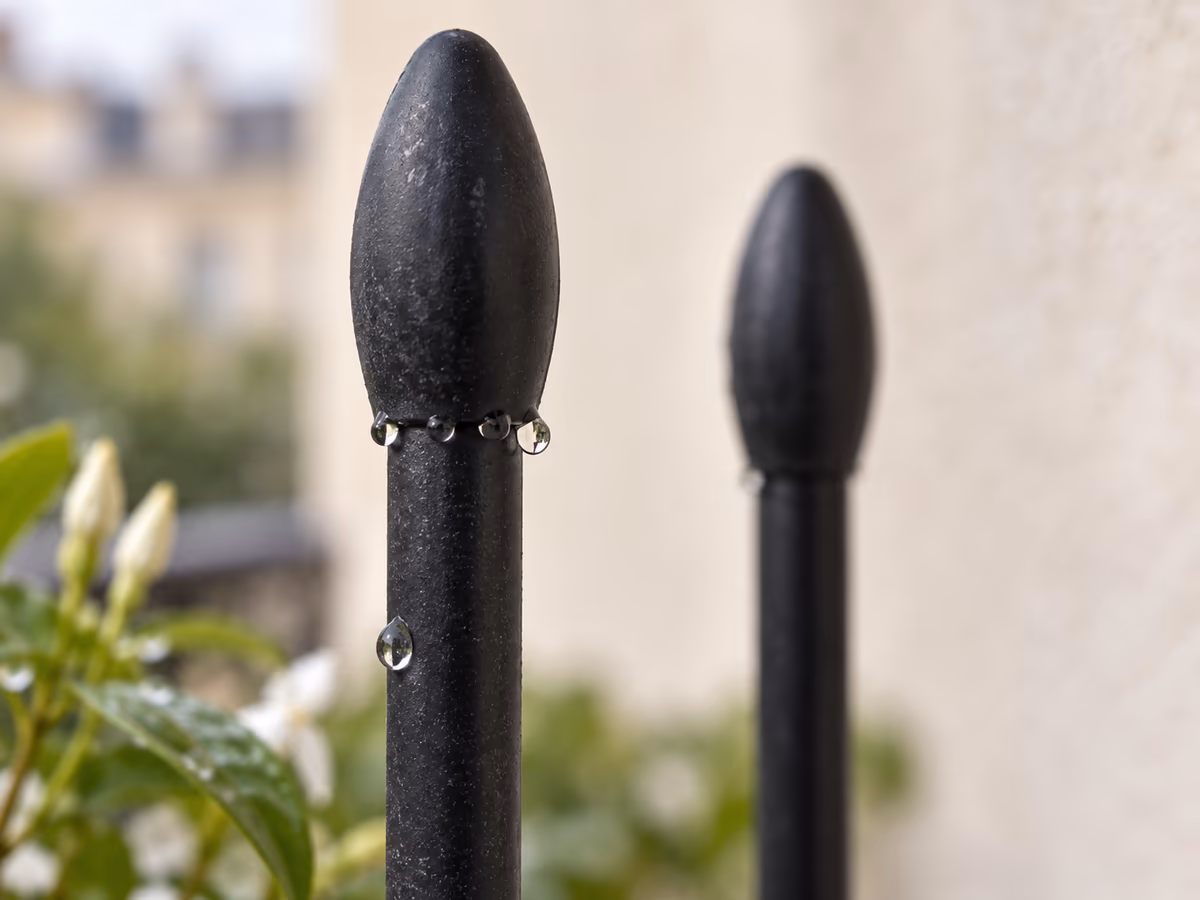 Gros plan du raccord entre un montant métallique noir et son embout plastique en haut d'un treillis de balcon, gouttes d'eau retenues au joint