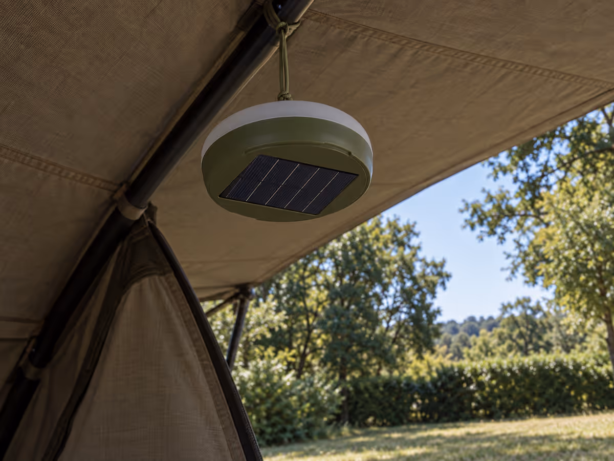 Boîtier solaire suspendu à l'ombre sous une toile de tente, panneau bleu visible au dos, arbres en arrière-plan