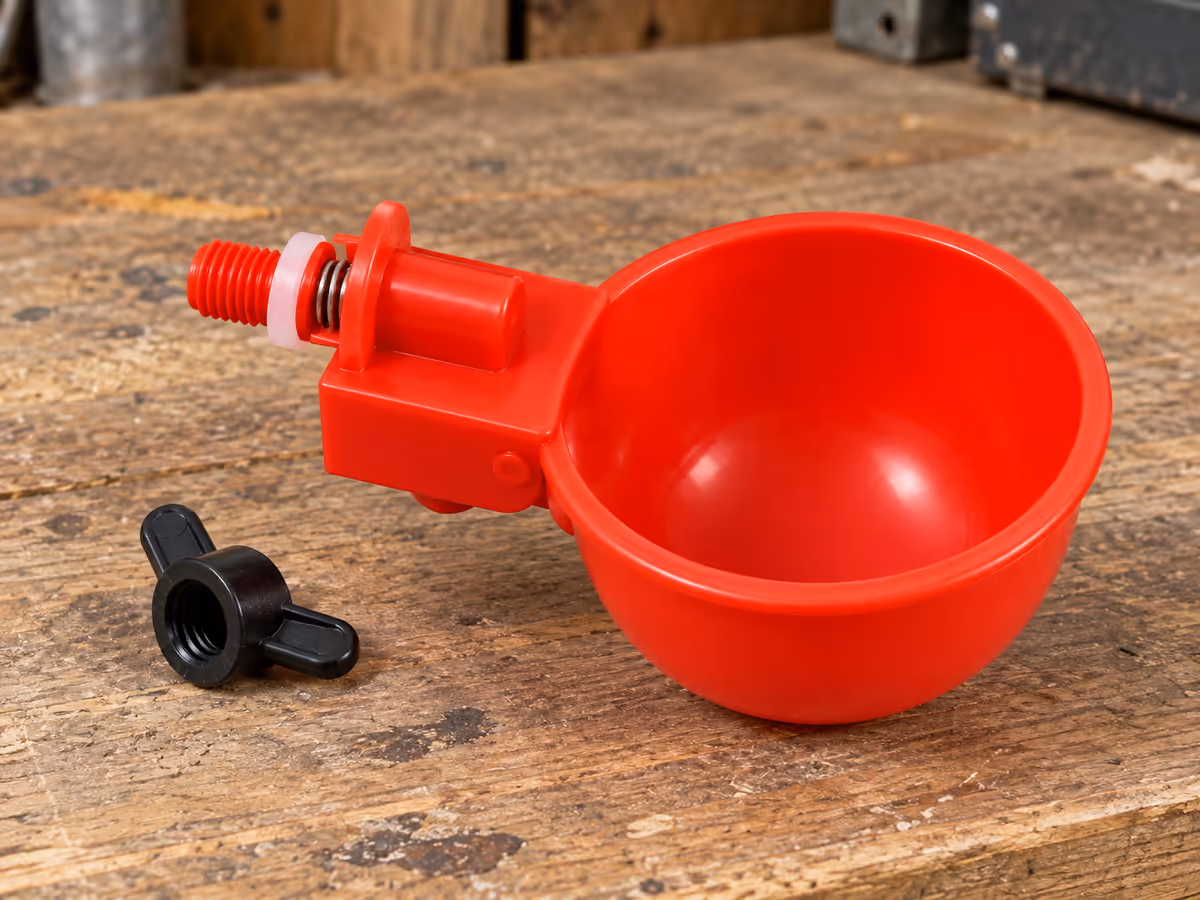 Bol abreuvoir rouge vu de profil sur un établi en bois, petit ressort visible entre la coupe et son boîtier, écrou papillon noir à côté