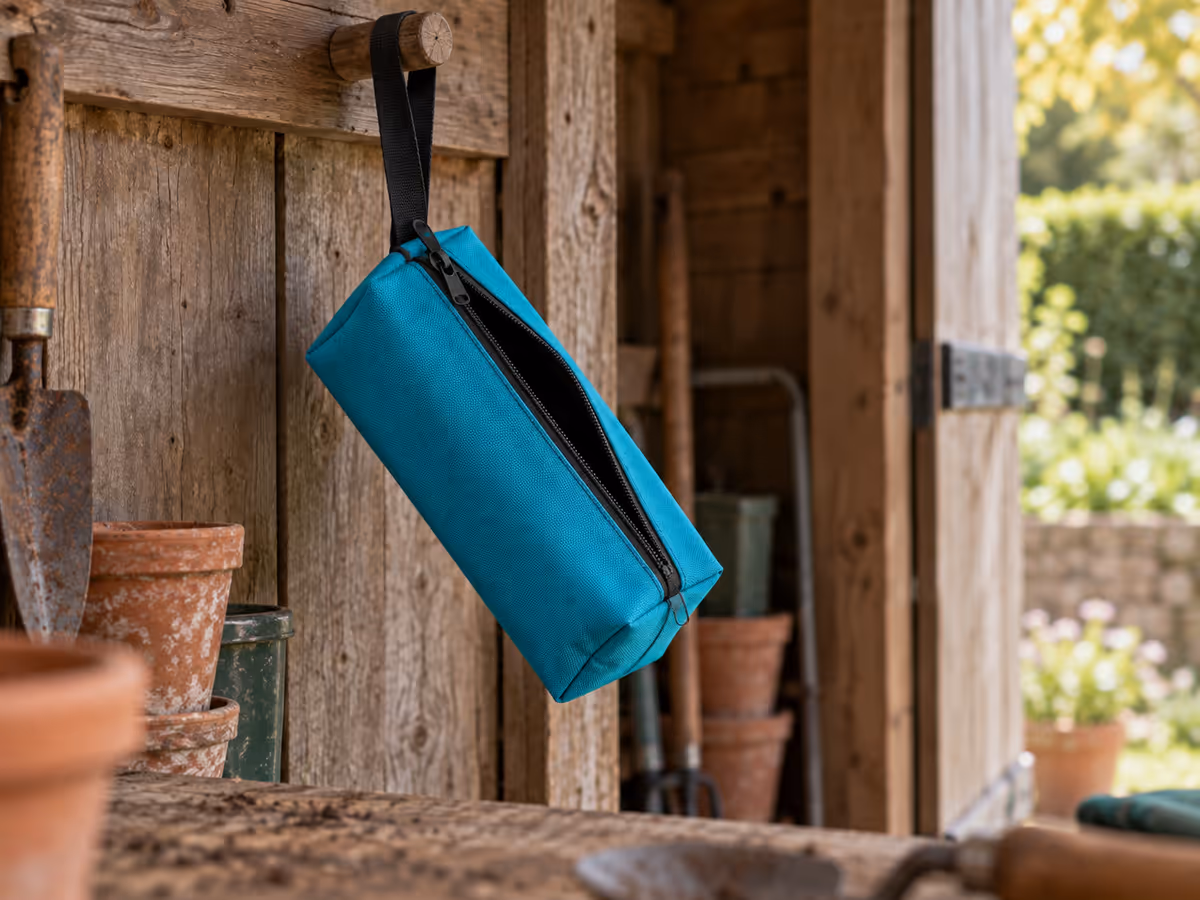 Pochette à outils en toile turquoise suspendue par sa sangle à un crochet de bois dans un abri de jardin, glissière entrouverte