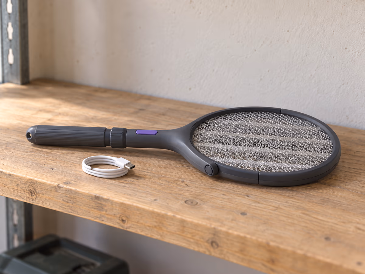 Raquette anti moustique pliée sur une étagère de garage, manche rétracté, câble USB-C enroulé à côté, lumière naturelle