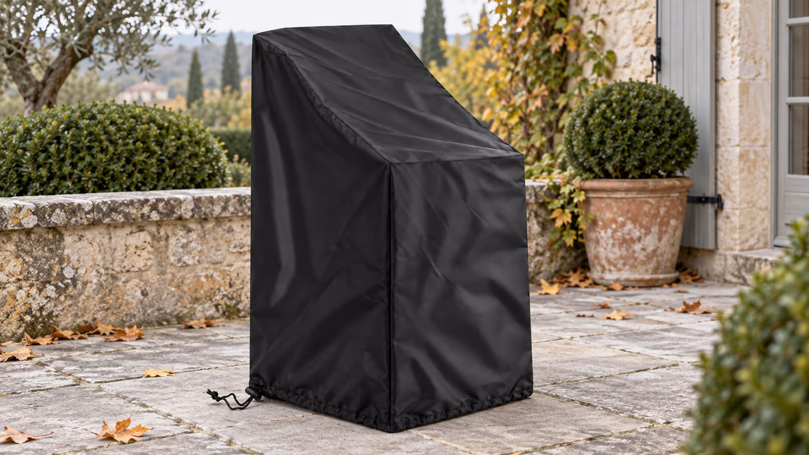 Housse de protection noire posée sur une pile de chaises de jardin empilables, cordon serré au pied, sur une terrasse en dalles en fin de saison