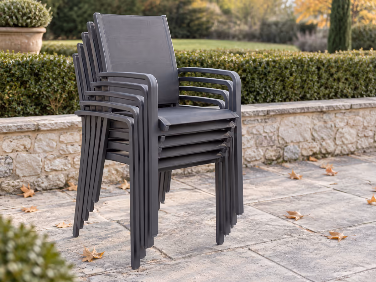 Pile de chaises de jardin empilées contre un mur de terrasse, dossiers alignés et assises emboîtées, sur dalles claires un matin sec d'automne